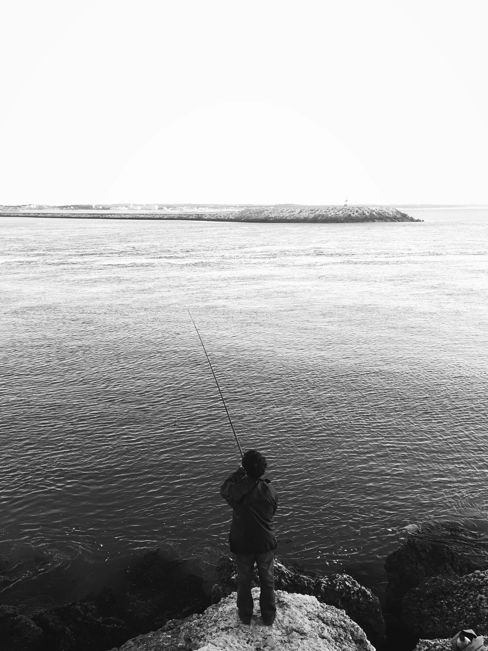 Person fishing from a rocky coastline in black and white