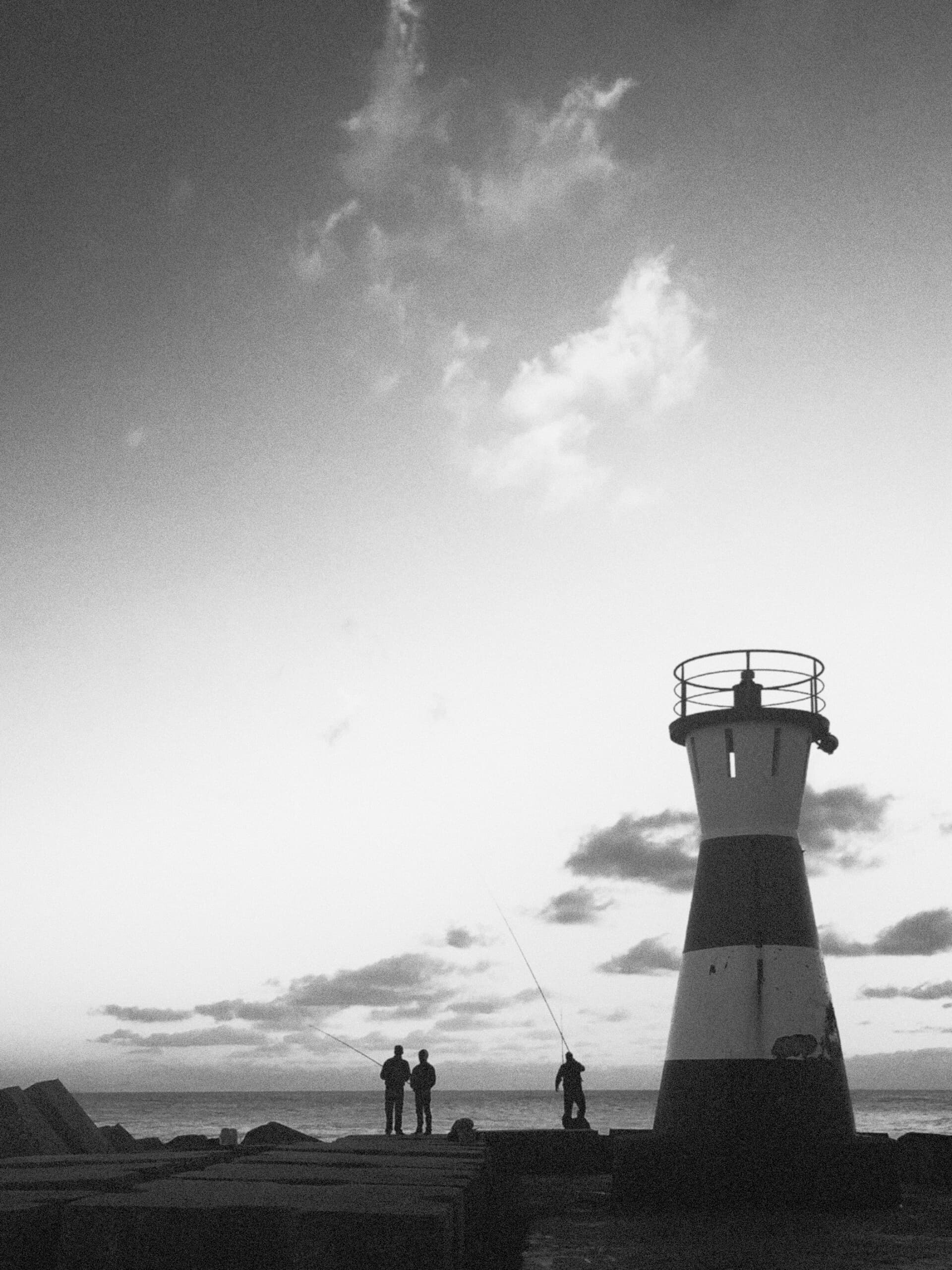 Fishermen silhouetted beside a lighthouse at dusk in black and white